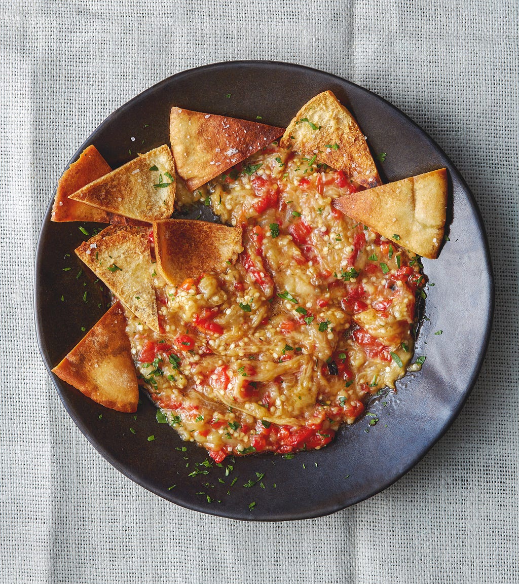 A wide dinner bowl of eggplant dip with toasted pita chips on it.