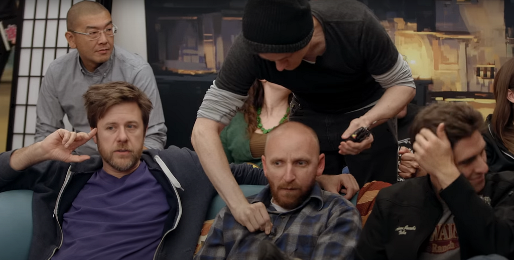Three Double Fine developers sit on a couch, with one more in the background. A 2 Player Productions documentarian wearing all black reaches from behind the couch to attach a lapel mic to the Double Fine employee in the center
