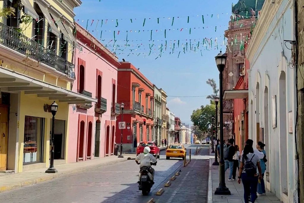 A street in Oaxaca with colorful buildings and people walking and riding scooters