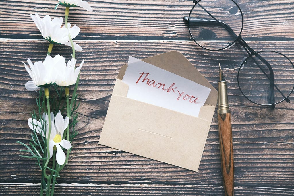 This image depicts white flowers, an opened letter with a note inside that peeks with the words “Thank you” written in red, a pen, and a glass with round frames on top of a wooden table.
