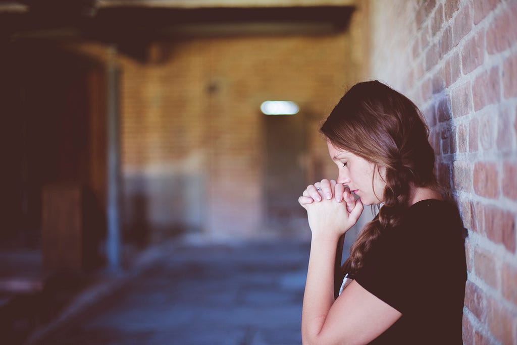 Woman with plaited braid praying with back to a brick wall.