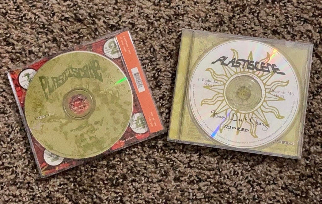 Two CDs in “J-card” plastic cases where labels are visible on brown/black carpet background— Plasticene EP left (yellow/green abstract design on CD with band name and a red/orange label behind it with a number in each corner), “Times Don’t Change” single right (yellow sun outline with band name top and single name on bottom on CD and yellow label underneath)