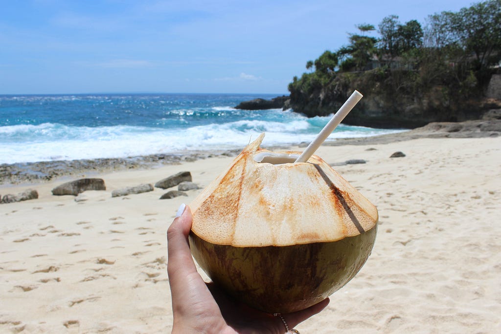 Person holding a coconut shell with a straw inside on the beach
