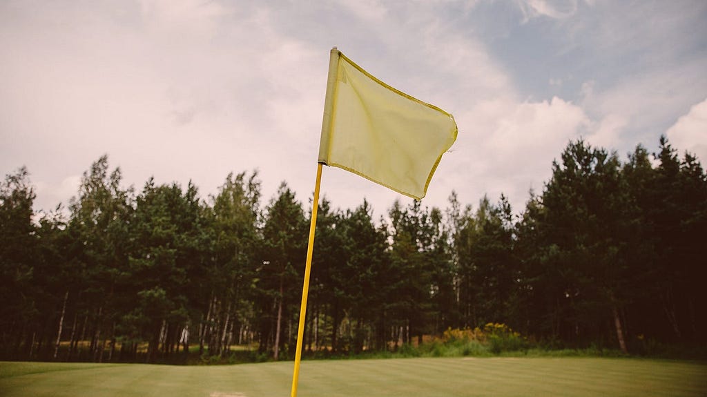 A green flag on a manicured golf course with trees lining an afternoon sky in the background.