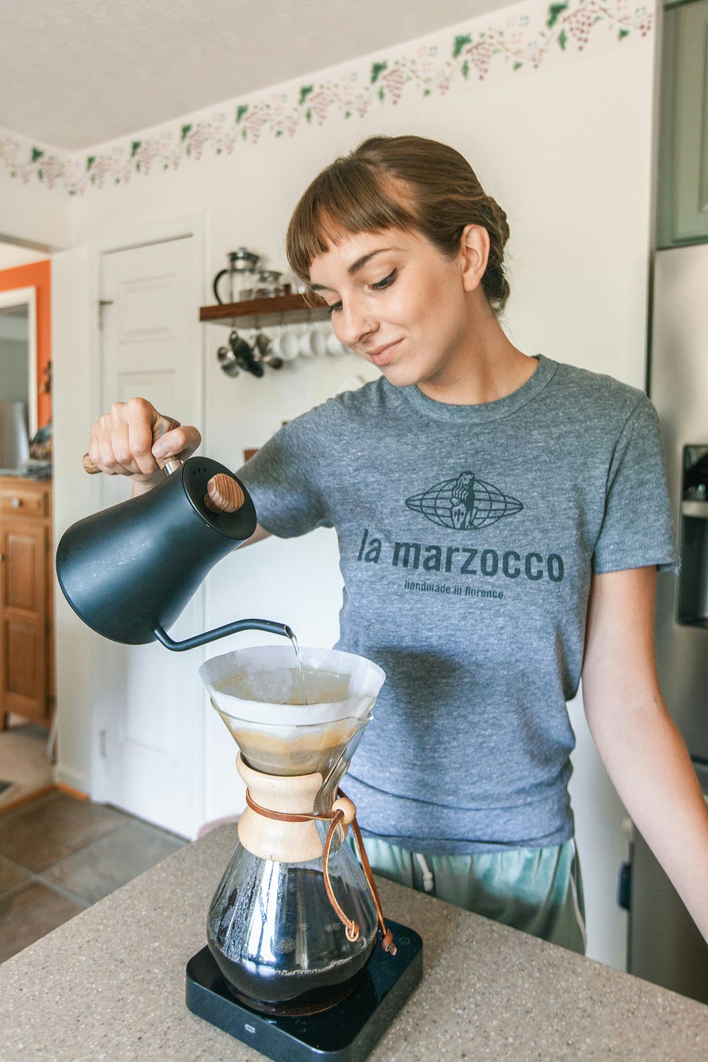 a young woman, happily adding warm water to her home brew coffee in her kitchen