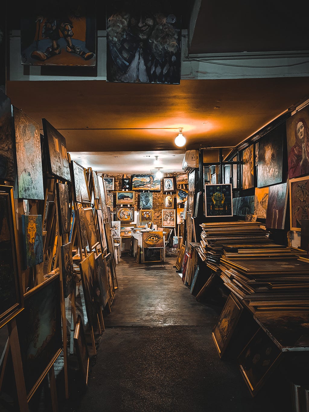 Overhead bare light on paintings all over walls and tables. Very little standing space. Warm tones of colour.