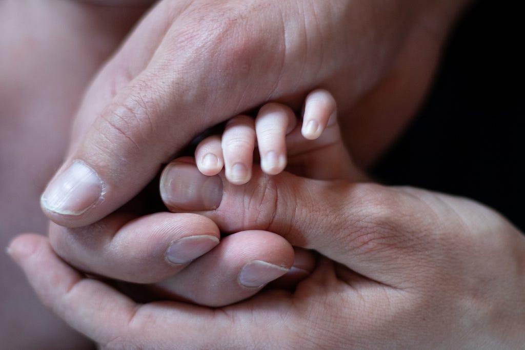 A picture of two adult hands holding a baby’s hand.