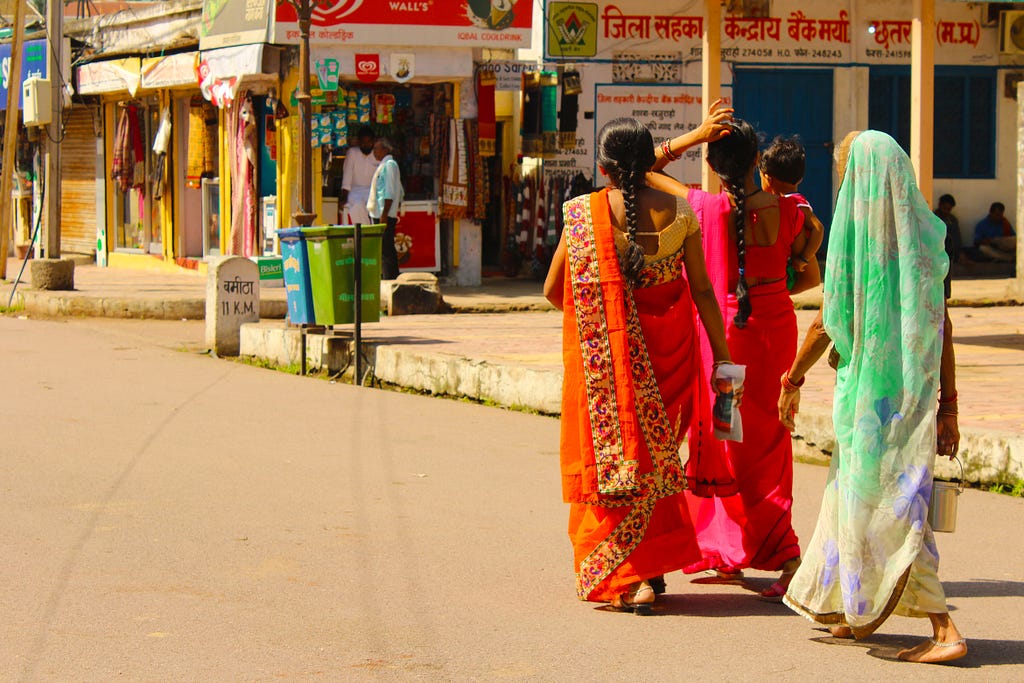 A group of three Indian women in saris walk dow the street at midday by shops with signs written in Hindi.