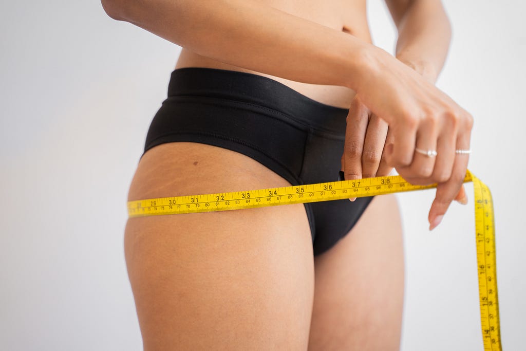 A woman in black underwear measuring herself around the hips with a yellow measuring tape.