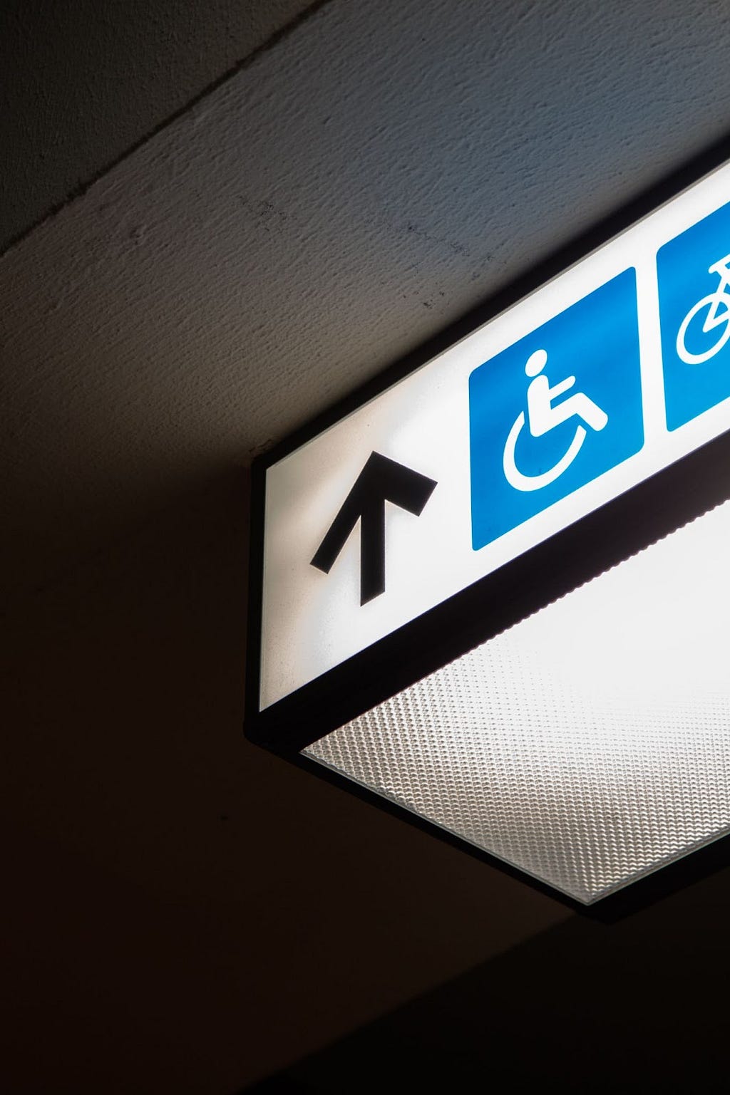 A photo of an illuminate white sign hanging from a ceiling. The front of the sign features a blue and white disability symbol of a person in a wheelchair, next to a black arrow pointing straight ahead.