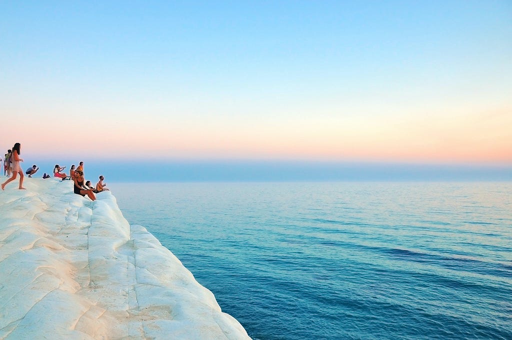 A group of people sitting on a large white rock looking out over a deep blue sea. The sky above is blue and the sun is creating a rainbow gradient effect as it sets on the horizon.