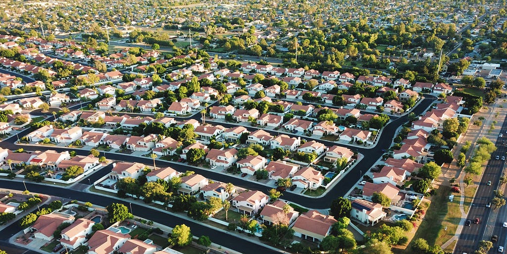 A bird’s-eye view of modern suburb. The houses have tiled roofs. There are multiple trees. Everything is tidy.