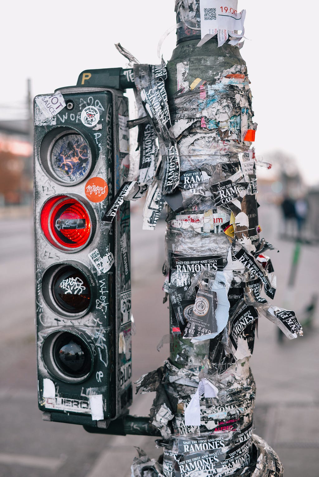 Bicycle traffic lights, red light is on for turning left. Light pole and light is covered with ramones stickers, signs and grafitti.