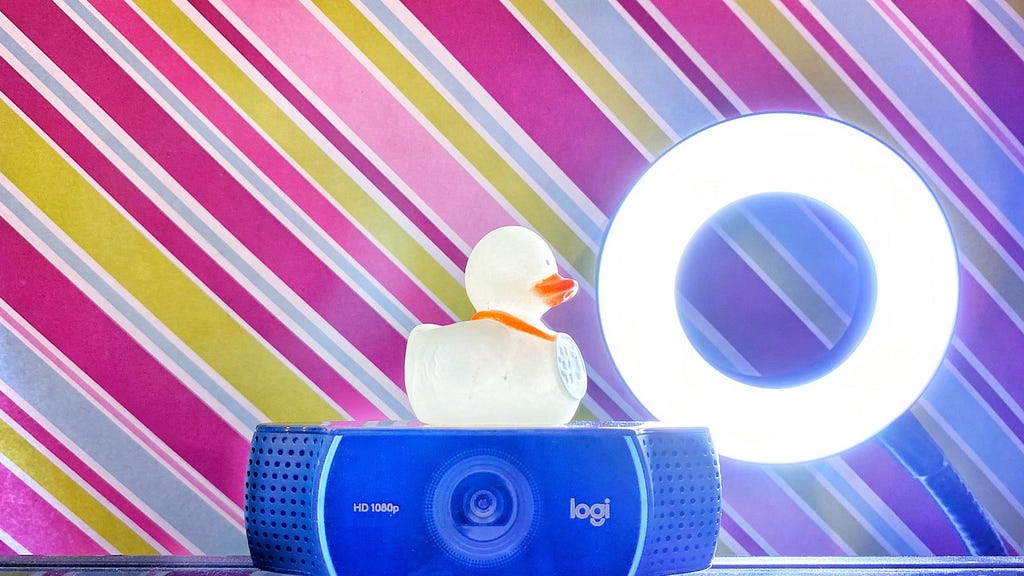 a small rubber duck on top of a web camera, with a ring light in the background and bright colors.