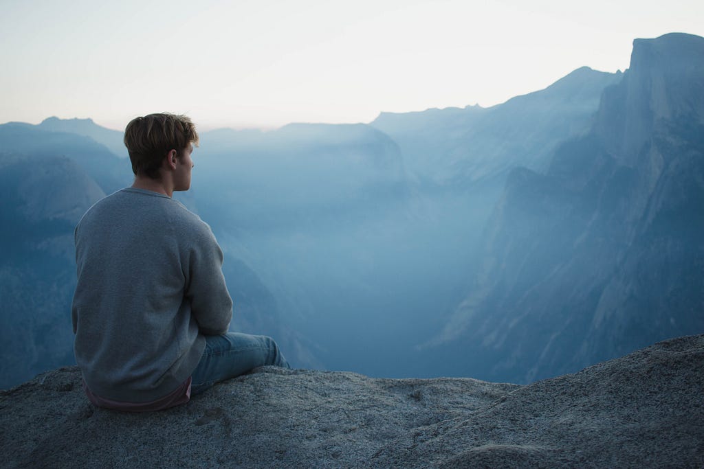 A person with short hair in a gray sweat shirt sitting on the edge of a ledge and looking thoughtfully into the distance full of foggy cliffs