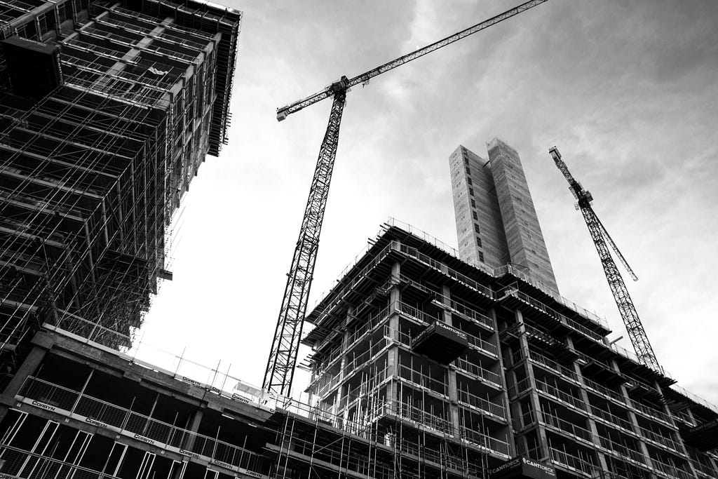 A black and white picture of a building under construction, with a tall crane in the center. The perspective is from below, highlighting the tallness of the crane.
