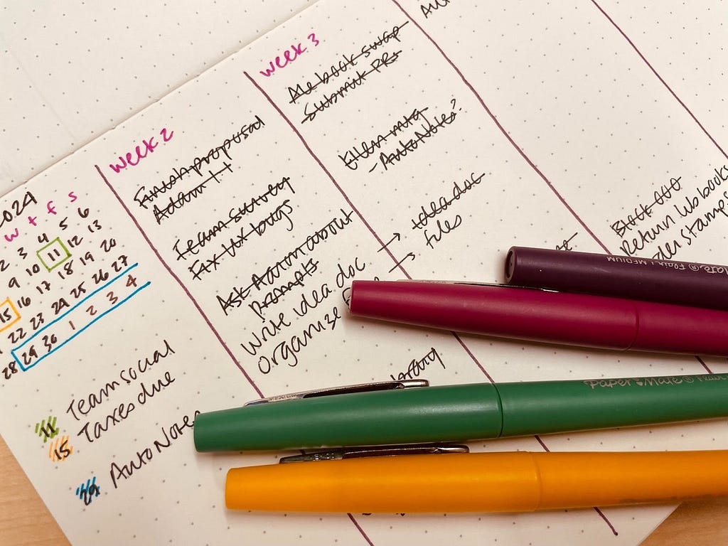 A photo of Vivian’s bullet journal, open to a page with a hand-drawn calendar and two handwritten vertical checklists labeled “Week 2” and “Week 3”; some checklist items are crossed out. The bullet journal page is partially obscured by four felt-tip markers that were used to write the page content.