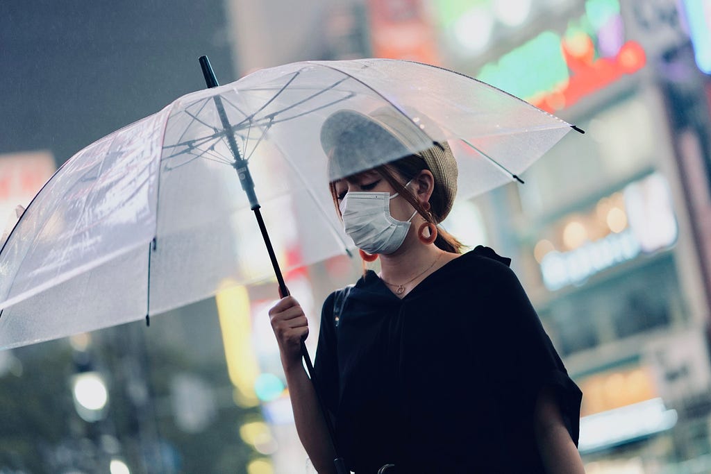 A Japanese woman wearing a medical mask and black blouse walks toward the left-center of the frame while carrying clear umbrella