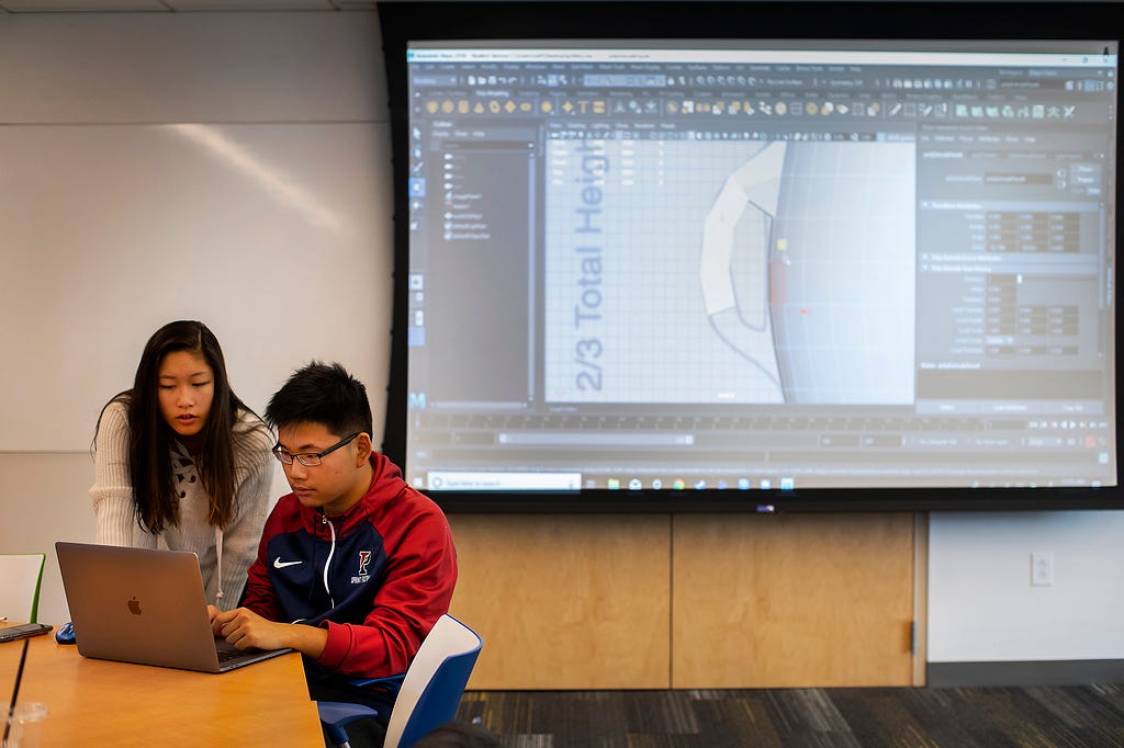 Two students work at a laptop. A screen behind them shows the 3D model of a jug they are designing.