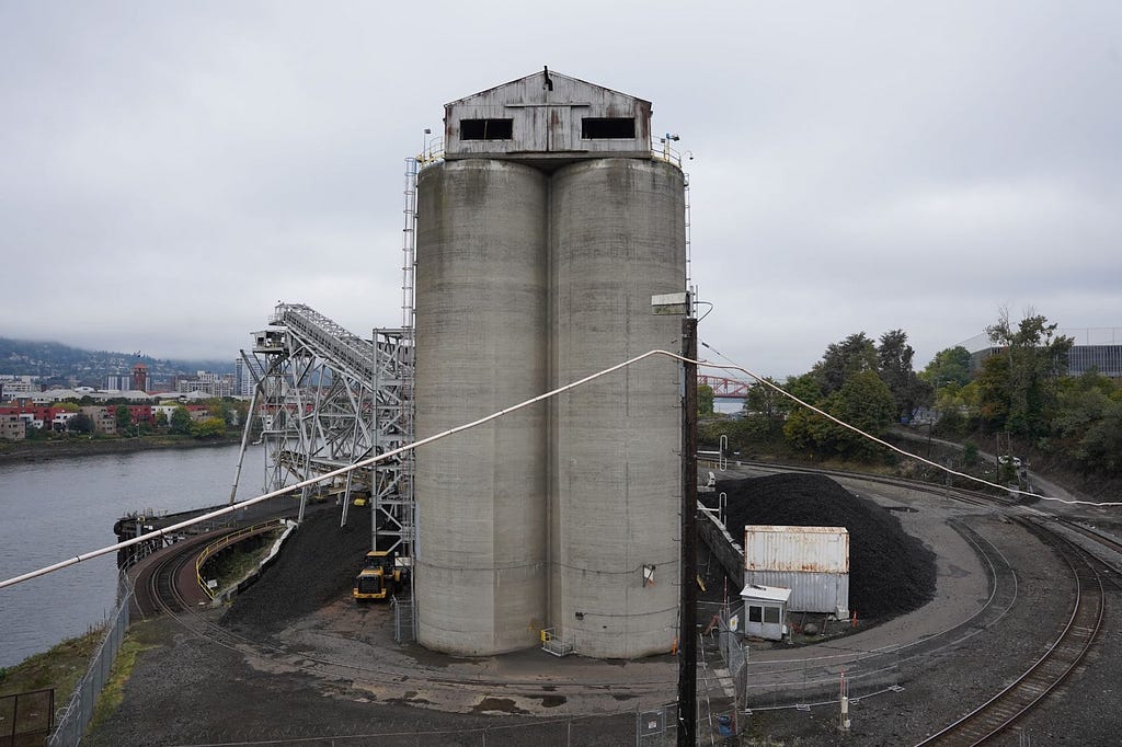 A photograph of an industrial lot with a large set of silos surrounded by train tracks. In the background, there is a river, trees, and buildings.