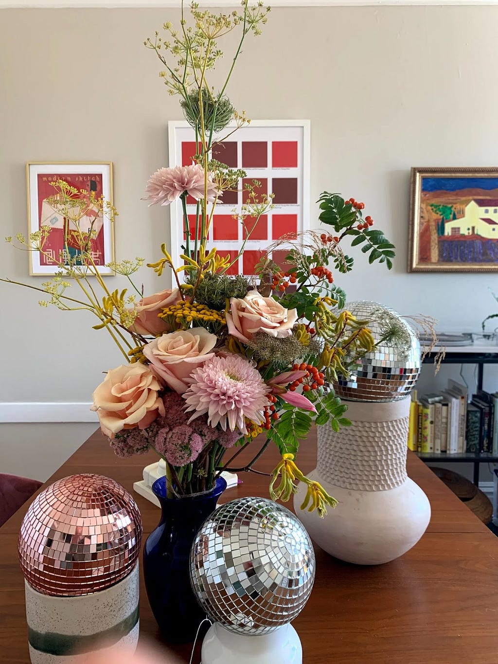 A large sunny bouquet of flowers, including pink roses and other pink and red flowers. The bouquet is on a table surrounded by small disco balls, and paintings are on the wall behind them.