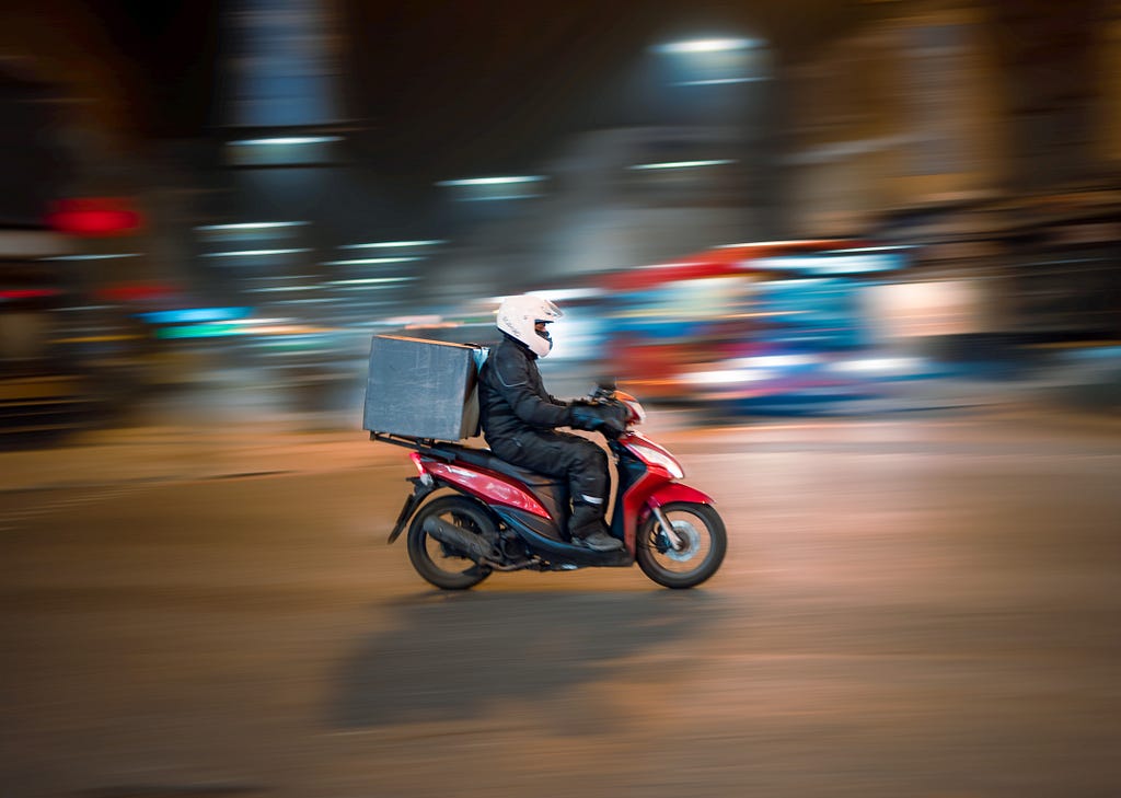 Homem com macacão preto pilotando uma motocicleta vermelha de entrega de delivery com fundo de cidade à noite desfocada.