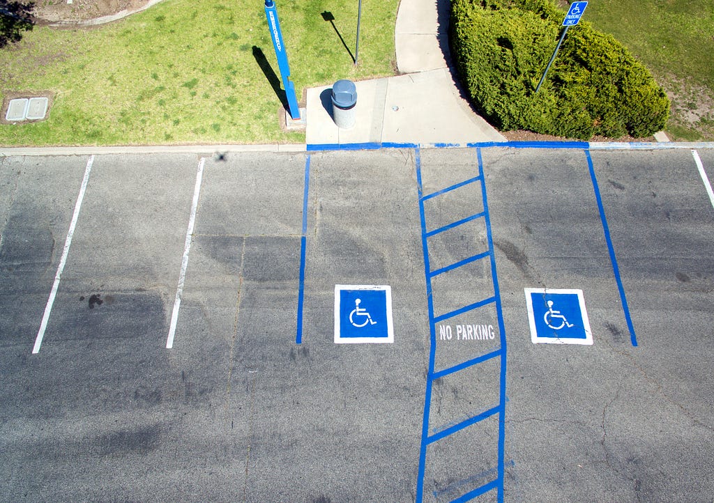 A zoomed-out image of two accessible parking spaces side-by-side, sharing an accessible aisle. The parking lot is grey, and the accessible parking spaces are painted in bright blue paint.