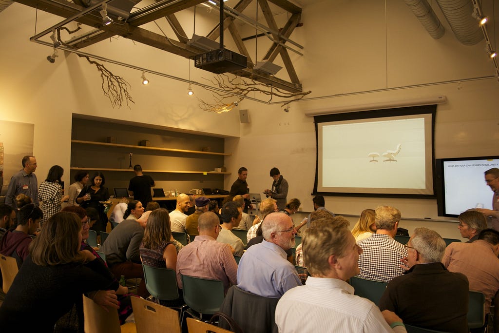 A room full of designers at IDEO waiting for a series of talks about design thinking and remote collaboration at scale to begin. There is a picture of an Eames chair projected on the screen, and the space is modern, clean, and creative.