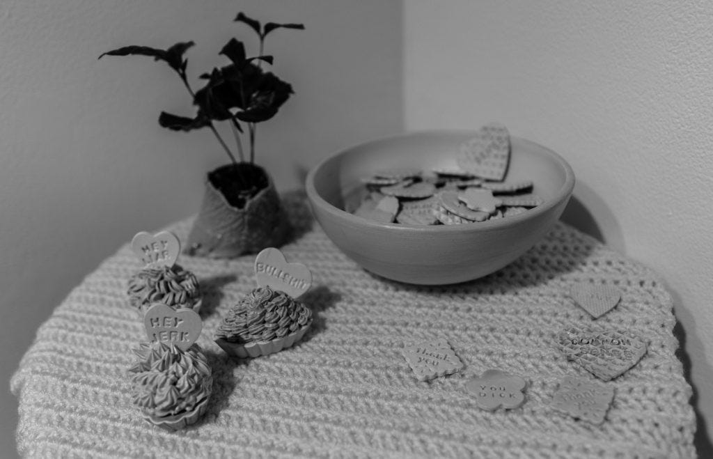 "Hey Liar." "Bullshit." "Hey Jerk." End table including ceramic custard cups with candy hearts made by Claudia Solorzano.