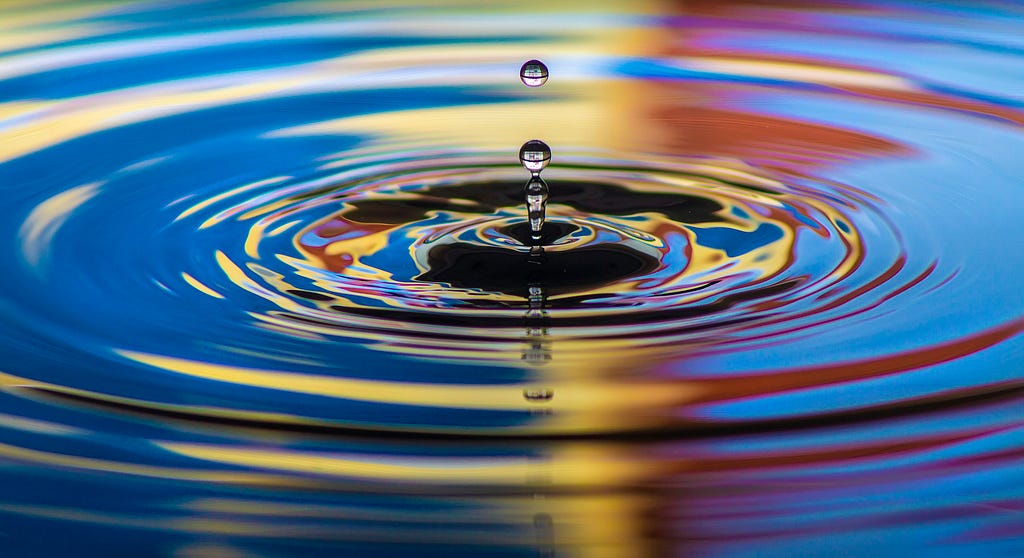 A close-up photograph of a water droplet falling into a pool, creating expanding circular ripples. The water reflects vivid colors — blue, yellow, red, and black — forming abstract patterns across the surface. The droplet is captured midair, perfectly centered above the rippling splash.