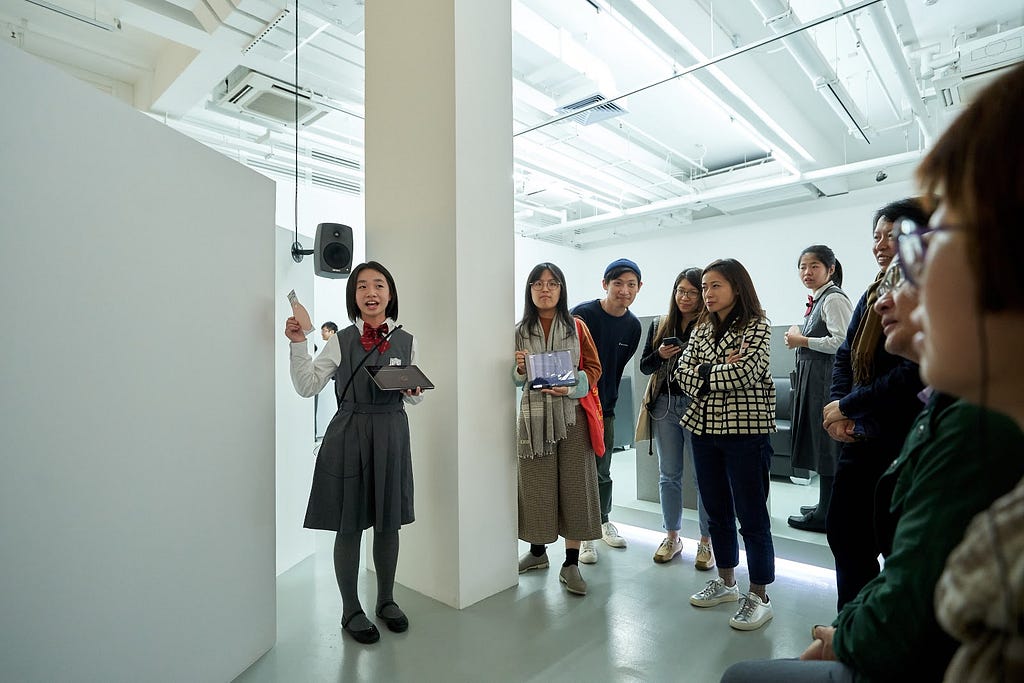A young student stands inside a gallery space. She speaks to a group of people that surrounds her.