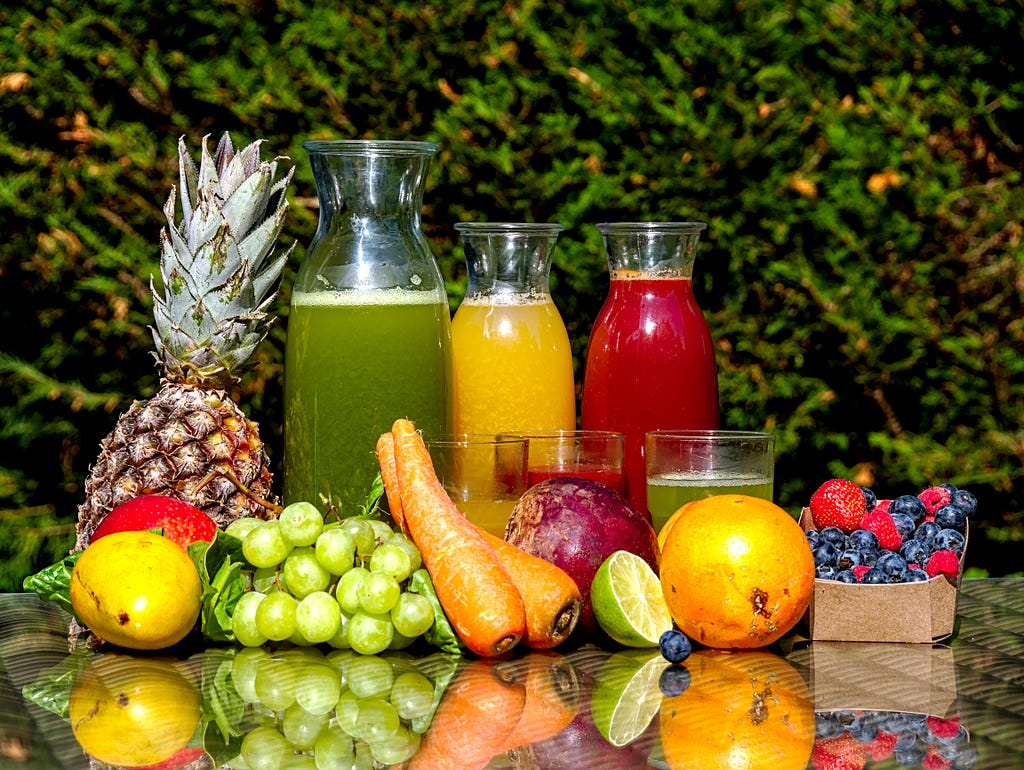 A set up of assorted fruits and what green, orange and beetroot juices and smoothies in transparent bottles.