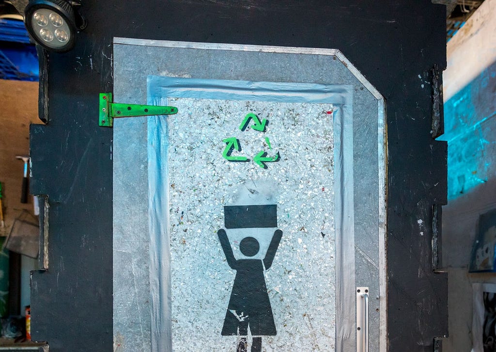 Outside of a portable toilet, made of recyclable materials. Color is silver and black with a graphic of a woman (indicating female toilet) and a bright green recycling symbol on front.