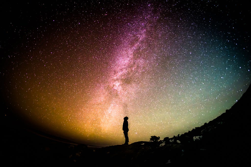 Photo of a rainbow, starry sky against a black background. The sky is visible in round form, taking up most of the image, with the curve towards the bottom of the image. Outside of the curved sky is black. In the centre of the image stands a person, a silhouette with neck bent, looking up towards the cosmos.