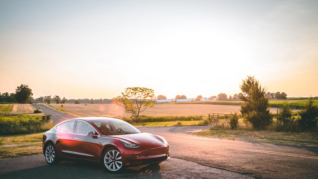 A red Tesla car infront of a flat field with a shining sun in the background