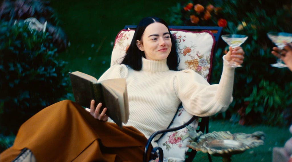 Emma Stone as Bella Baxter in Poor things, sitting on a chair and toasting a martini whilst reading a book