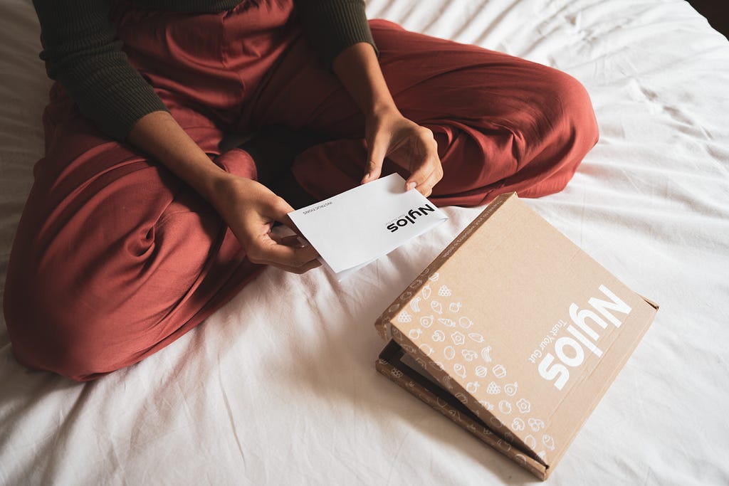 A person sits on a bed and holds a manual in her hands. A cardboard box lies on the bed in front of her.