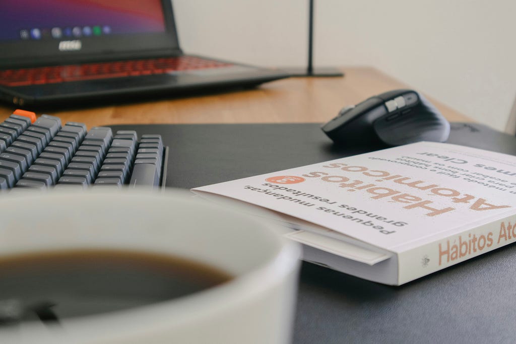 Foto de uma mesa de trabalho, com um computador e um teclado ao fundo. Em destaque, uma caneca de café e o livro Hábitos Atômicos, de James Clear.