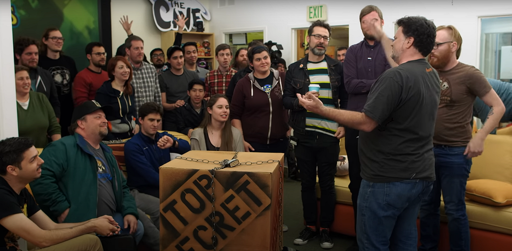 Dozens of smiling Double Fine employees crowd around CEO Tim Schafer in a meeting room. To Shafer’s left is a cardboard box marked “top secret,” and tied closed with a chain. This is an opening ceremony for Amnesia Fortnight, a Double Fine company game jam