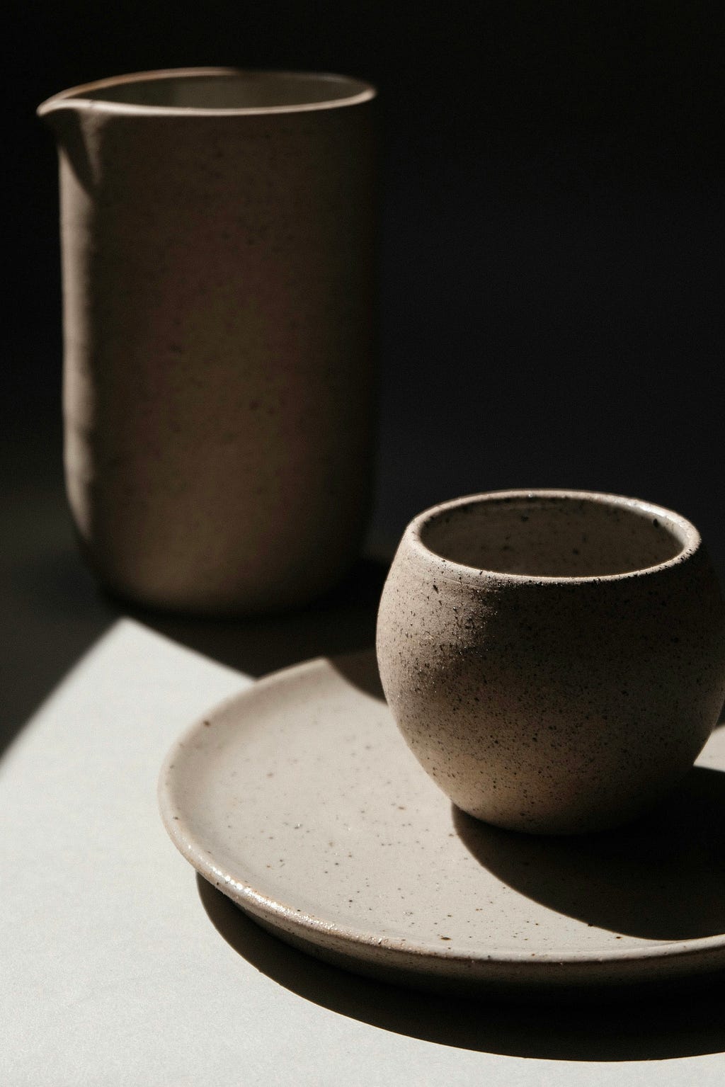 Three stoneware articles — plate, cup, and jug — stand on a surface. The stoneware articles and surface are partly lit by sunlight.