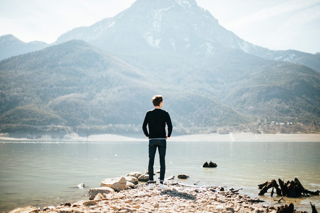A man stands alone on a spit of land jutting into a calm lake as a mountain silently observes from the background. The place is serene, yet remarkably, palpably, lonely.