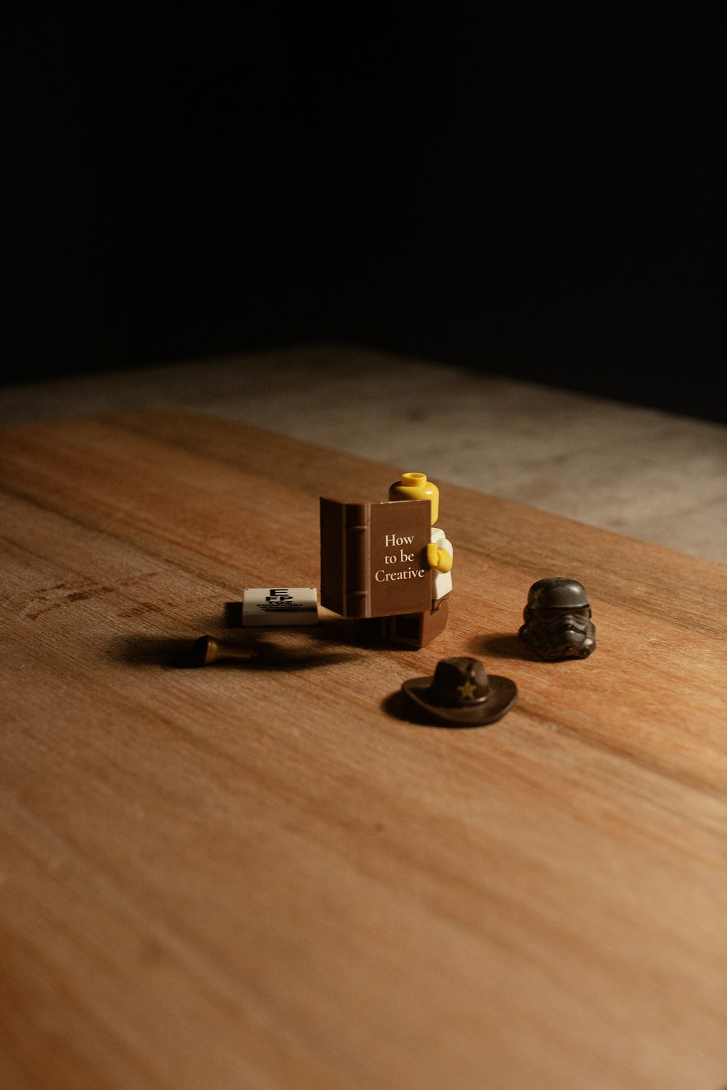 A brown table top with a Lego figure holding a brown book with the words “How to be Creative”.