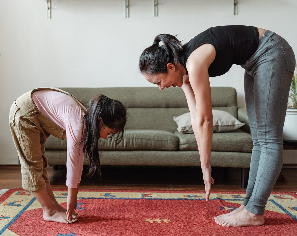 A mother and daughter stretch together, standing and touching their toes, in front of a sofa.