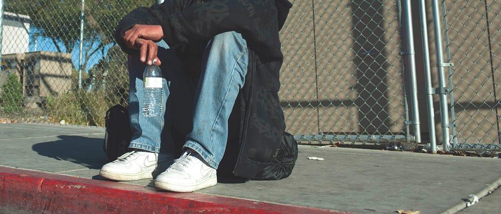 adult sitting on curb holding water bottle wearing jeans, black hoodie, and white sneakers