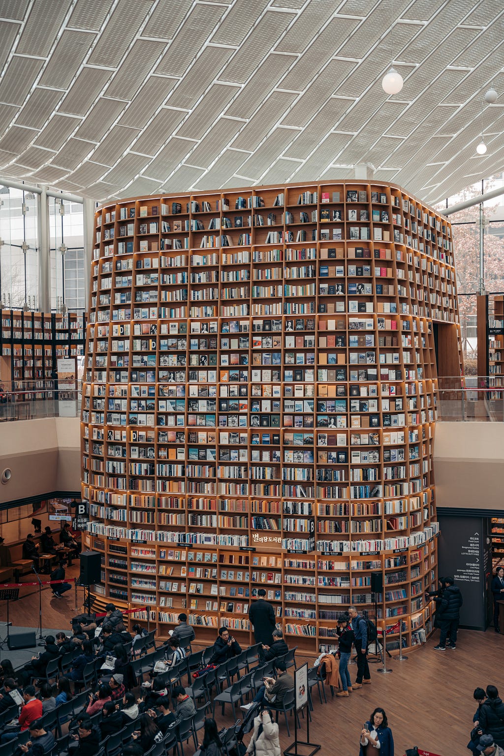 a group of people in the foreground, some sitting in chairs in a row, some moving around, in a library or bookstore with a two-story cube shaped book case taking up most of the space