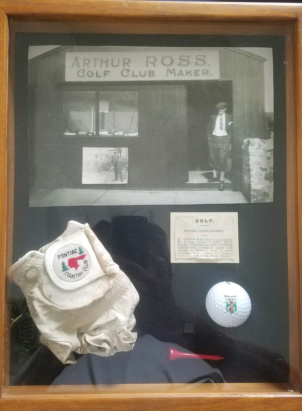 A framed picture put together by my dad containing various golf items. At the top is a full picture of Arthur Ross in front of his golf club shop in Carnoustie, Scotland. There is a small newspaper article announcing his opening of the shop. My dad added his golf glove, a ball, and a tee. From the family archives, photo by author.