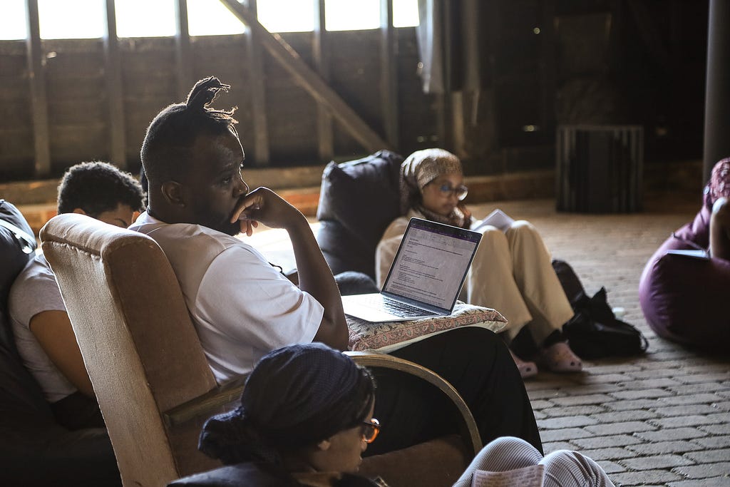 The group sit around during the Fireside Chat. Guest Junior Muya from Government Digital Services looks on as he listens. Three other guests are in view.