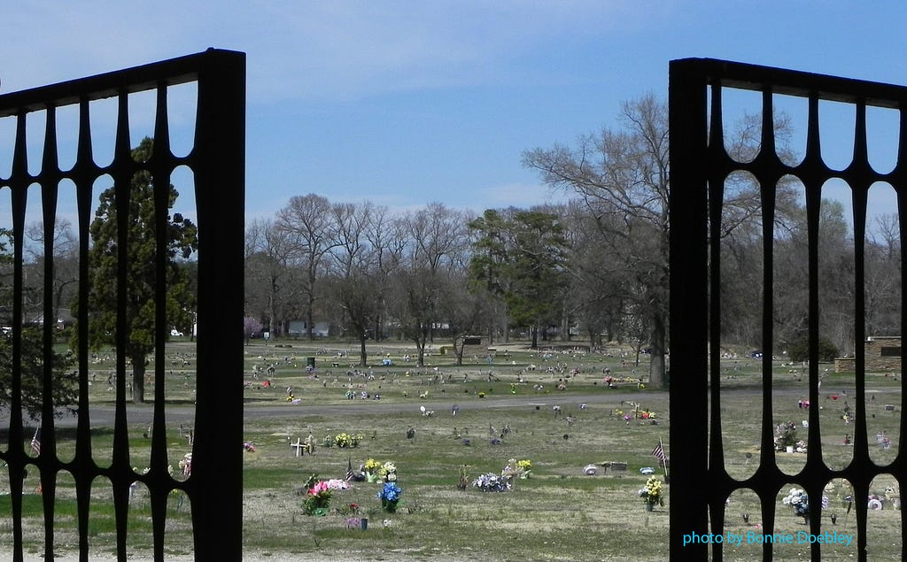 View of a cemetery from the open gates with many graves under a blue sky. Many of the graves are decorated with urns of flowers.