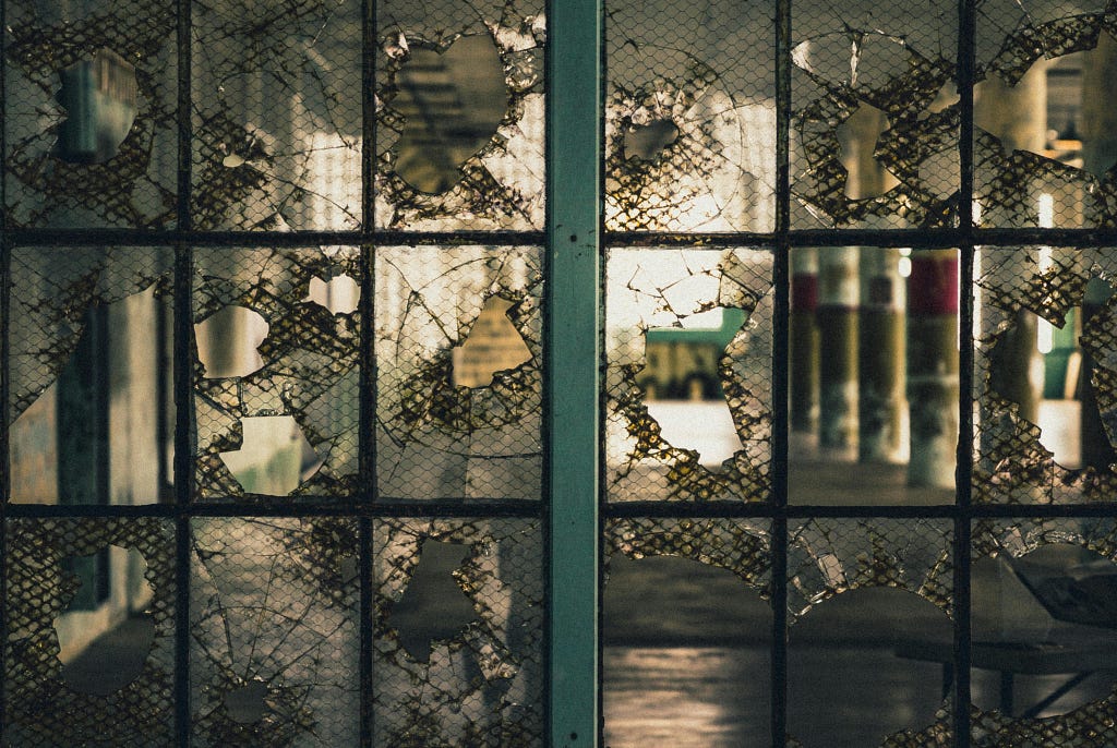 Close-up of a shattered wire-reinforced glass window. The broken panes reveal a dim interior with reflections and distorted shapes, suggesting fragmentation, institutional coldness, and emotional rupture.