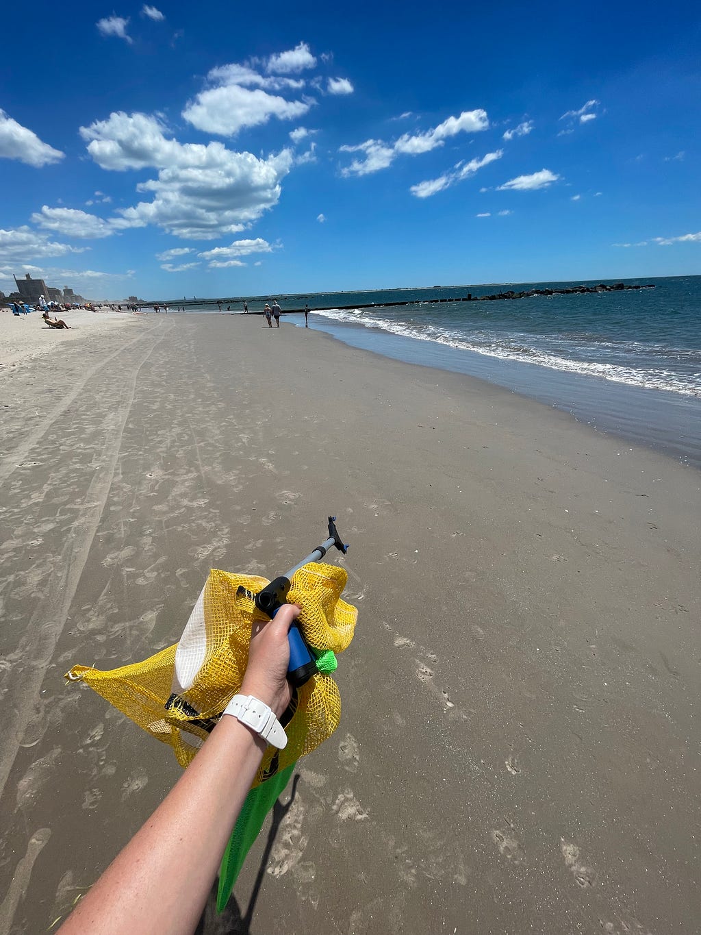 An aerial shot of my left hand holding a trash picker and reusable bags as I walk down Coney Island Beach with the Atlantic Ocean on my right and beachgoers in the foreground